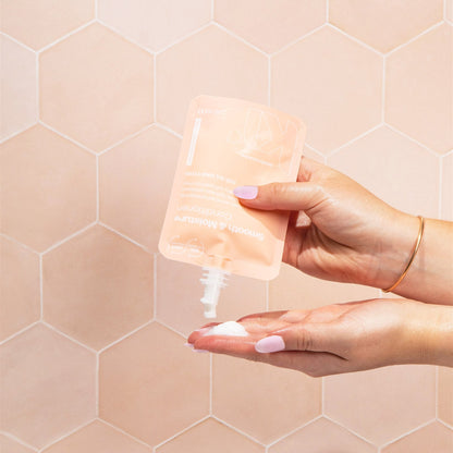 Hand holding a small bottle of cream against a pink tiled wall.