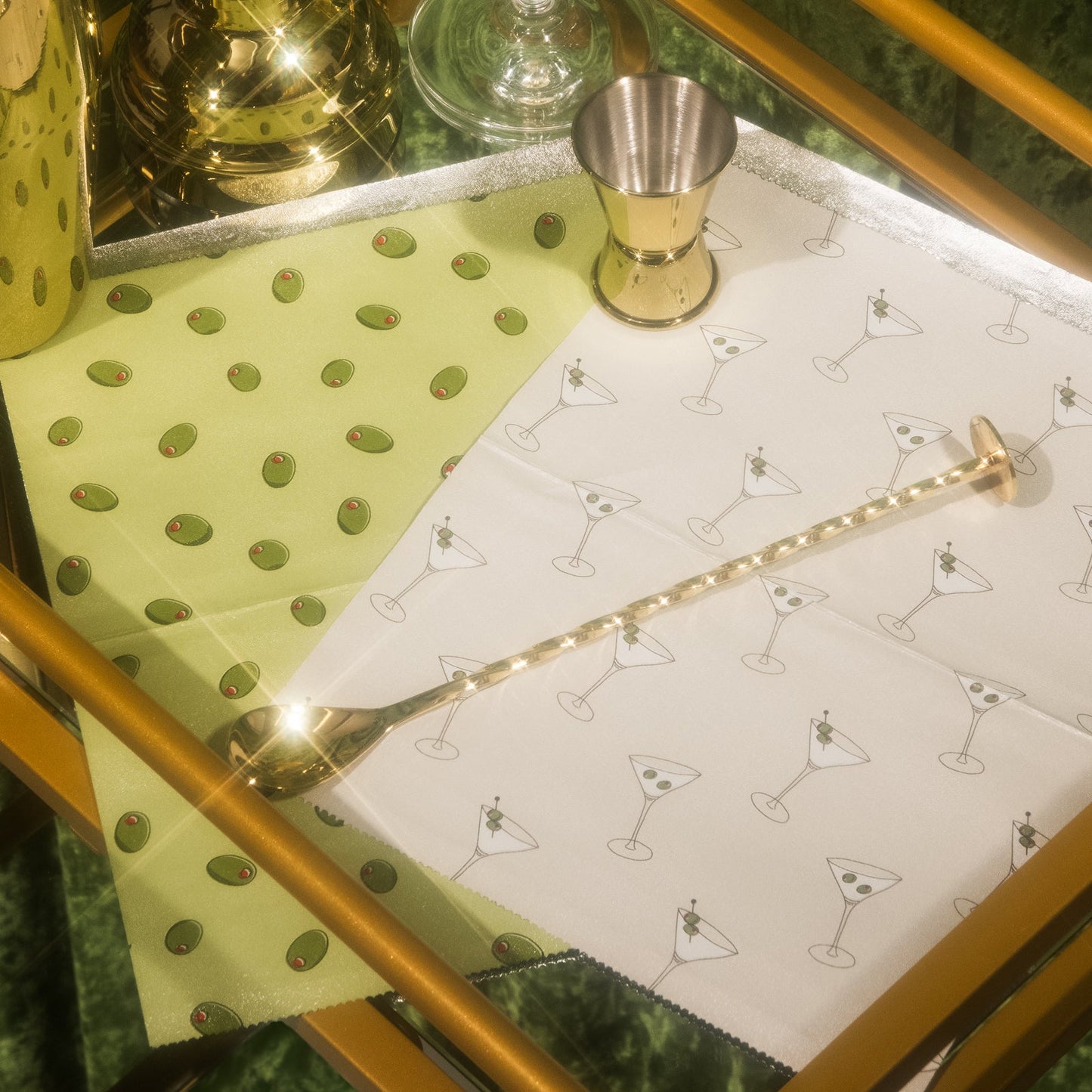 Bar cart with a green polka dot napkin, white napkin with cocktail glass pattern, and gold bar spoon.