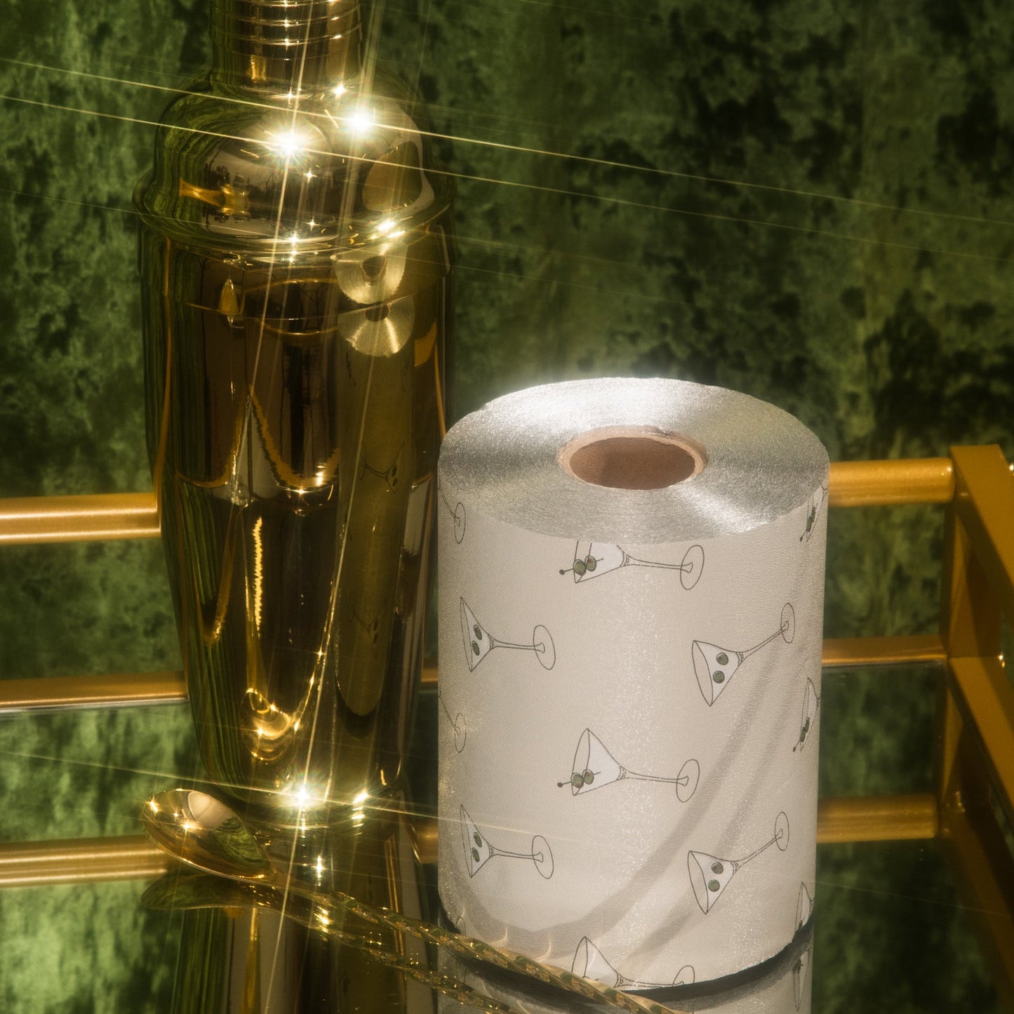 Gold cocktail shaker next to a roll of paper towels with cocktail-themed design on a green marble surface.
